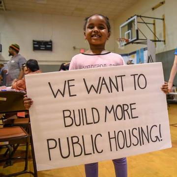 Child holds a sign saying 'we want more public housing'