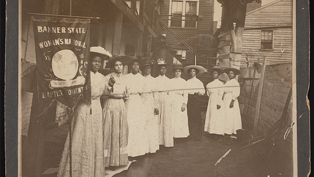A group of women in turn-of-the-19th-century clothing stand next to a banner with indistinguishable text