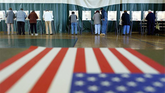 People stand in voting booths, with the American flag in the foreground