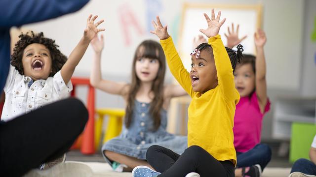 Several preschool age children sit on the floor and raise their arms