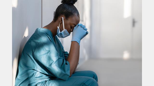 A female health care worker in scrubs and a mask, resting against a wall