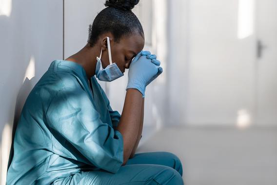 A female health care worker in a mask and scrubs, leaning against a wall