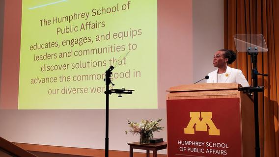 Dean Nisha Botchwey stands onstage next to a slide printed with the Humphrey School mission statement